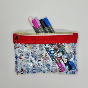 May include: Clear plastic pencil case with a red zipper and a nautical themed print. The print features lighthouses, anchors, boats, and the text "The Beach is Good". The case is filled with four colorful markers.