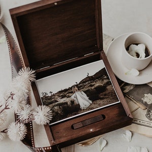 May include: A wooden box with a hinged lid, open to reveal a photograph of a person in a white dress standing in a desert landscape. The box is sitting on a table with a cup of coffee and a newspaper.