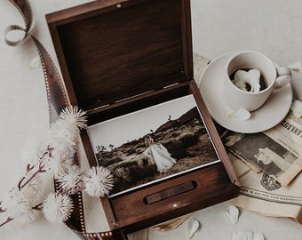 May include: A wooden box with a hinged lid, open to reveal a photograph of a person in a white dress standing in a desert landscape. The box is sitting on a table with a cup of coffee and a newspaper.