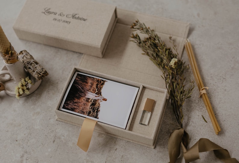 May include: A beige linen box with a gold ribbon and a photo of a person in a field. The box also contains a small gold USB drive. The box is labeled "Laura & Antoine 02.27.2022"