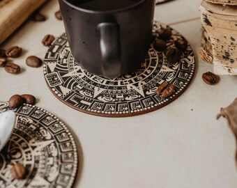 May include: A black ceramic mug filled with a dark liquid sits on a round coaster with a black and tan Aztec calendar design. Coffee beans are scattered around the mug and coasters. Additional coasters and coffee beans are visible in the background.