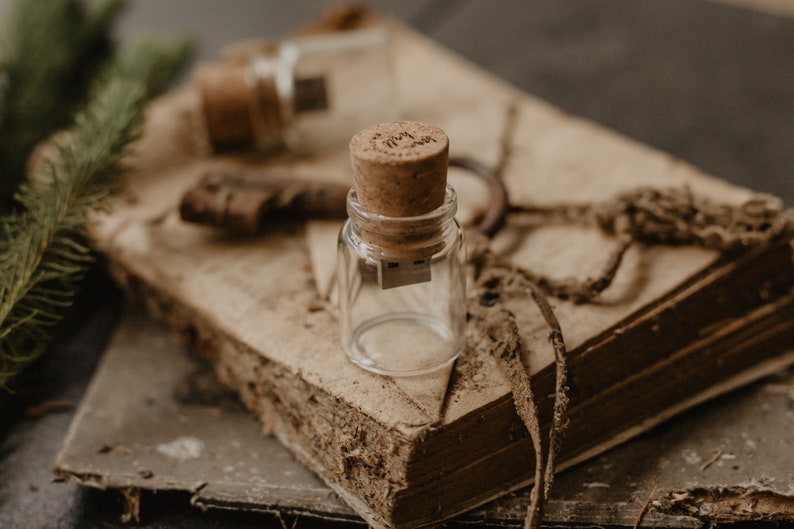 May include: A small glass bottle with a cork top and a USB drive inside. The bottle is sitting on an old, worn book with a brown leather cover.
