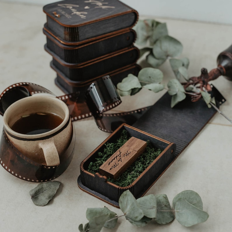 Peut inclure: Une pile de bo&icirc;tes en bois sombre avec du texte grav&eacute;, une tasse en c&eacute;ramique entour&eacute;e d'une bande de film 35mm, et une bo&icirc;te en bois ouverte contenant une cl&eacute; USB en bois nich&eacute;e dans de la mousse verte, agr&eacute;ment&eacute;e de feuilles d'eucalyptus.