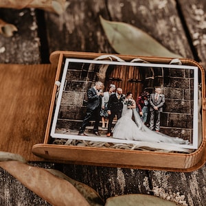 Pode incluir: Uma caixa retangular de madeira com tampa articulada contendo uma fotografia impressa de um grupo de casamento em frente a um edifício de pedra. A caixa é feita de madeira castanha clara e apresenta um acabamento liso.
