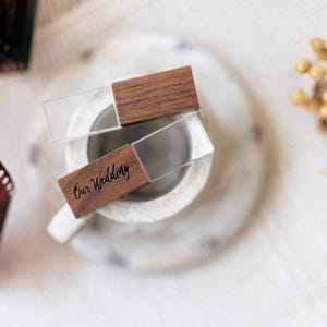 Könnte beinhalten: Zwei rechteckige USB-Sticks aus einer Kombination von klarem Acryl und dunkelbraunem Holz. Einer der Sticks ist mit dem Text "Our Wedding" in einer schwarzen Schreibschrift graviert. Die Sticks liegen auf einer kleinen, runden, cremeweißen Schale.