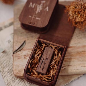 Könnte beinhalten: Ein dunkelbrauner USB-Stick aus Holz mit der Gravur Mr. & Mrs. Adamo auf der Oberfläche, eingebettet in eine passende Holzschachtel mit Papierstreifen. Der Deckel der Schachtel zeigt die Buchstaben M und A mit einem Blumenmotiv und einem Datum.