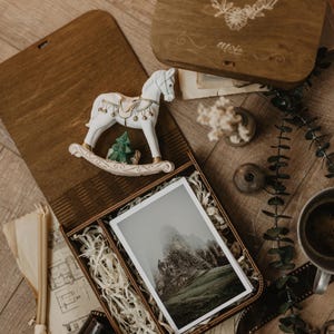 K&ouml;nnte beinhalten: Eine h&ouml;lzerne Erinnerungsbox mit einem Klappdeckel, der mit den Namen Veronica e Pietro und einem Blumenkranz graviert ist. Darin befinden sich eine wei&szlig;e Schaukelpferd-Figur, ein kleiner gr&uuml;ner Baum und ein gerahmtes Foto einer nebligen Berglandschaft.