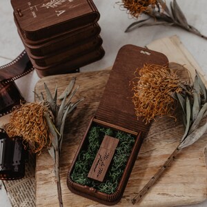 May include: A wooden box with a hinged lid, containing a small wooden USB drive with the inscription "Mr. & Mrs. Adamo" and a bed of green moss. The box is open and sitting on a wooden surface with a stack of other wooden boxes and dried flowers.