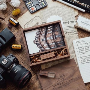 May include: A dark wood photo box containing a printed photograph of a building, with a matching wooden lid engraved with the names Gabby & Paul and the date 2021.03.06, accompanied by a wooden USB drive and wood shavings inside the box.