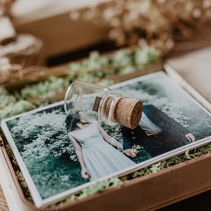 May include: A wooden box with a moss lining contains a printed photograph of a couple and a glass vial with a cork stopper. The vial contains a USB drive.