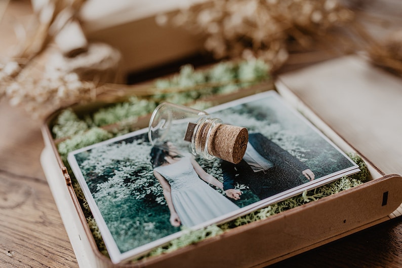 May include: A wooden box with a moss lining contains a printed photograph of a couple and a glass vial with a cork stopper. The vial contains a USB drive.