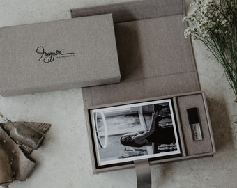 May include: A gray linen box with a black logo that says "Juggori Photography". The box is open and contains a black and white photograph of a woman in a dress, a small gray box with a USB drive, and a gray linen interior.