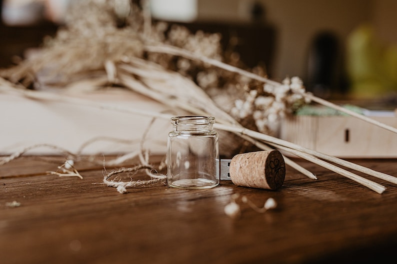 May include: A wooden USB drive shaped like a bottle with a cork top. The drive is sitting on a wooden table with dried flowers and twine.