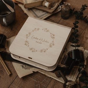 K&ouml;nnte beinhalten: Eine quadratische, helle Holzschachtel mit Klappdeckel, die mit einem lasergravierten Blumenkranz-Design verziert ist, welches den Text Emeline et Nicolas und das Datum 14.08.2024 umschlie&szlig;t. Die Schachtel hat abgerundete Ecken und einen Metallverschluss.