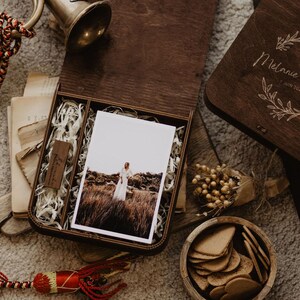 K&ouml;nnte beinhalten: Eine dunkelbraune Holzschatulle mit einem Foto einer Person in einem wei&szlig;en Kleid auf einem Feld, einem kleinen USB-Stick aus Holz und Papierf&uuml;llmaterial. Der Deckel ist mit einem Blumenkranz und dem Text Melanie & Viktor 7 JUIN 2024 graviert.