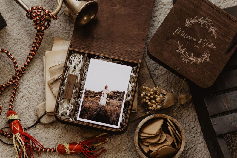 K&ouml;nnte beinhalten: Eine dunkelbraune Holzschatulle mit einem Foto einer Person in einem wei&szlig;en Kleid auf einem Feld, einem kleinen USB-Stick aus Holz und Papierf&uuml;llmaterial. Der Deckel ist mit einem Blumenkranz und dem Text Melanie & Viktor 7 JUIN 2024 graviert.