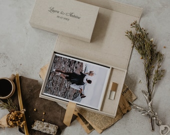 May include: A beige linen box with a photo album inside. The album contains a black and white photo of a couple walking on a beach. The box also includes a USB drive and a small piece of paper with the text "Laura & Antoine 22.07.2023" printed on it.