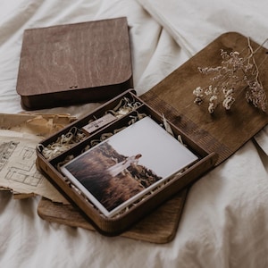 Pode incluir: Uma caixa de madeira castanho-escura com tampa articulada, contendo uma impressão fotográfica retangular de uma pessoa com um vestido branco num campo, uma pequena pen USB de madeira e flores secas, acompanhada por papéis antigos rasgados.