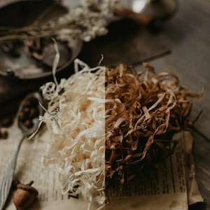 Puede incluir: Una composición de naturaleza muerta con dos mitades de papel triturado, una crema y otra marrón, sobre papel vintage. Una cuchara con granos de pimienta, una bellota y otros elementos decorativos completan la estética rústica.