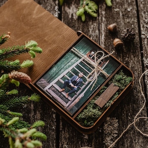 May include: A rectangular wooden box with a hinged lid containing a photograph of a person sitting on wooden stairs, a small wooden USB drive, and green moss. The photograph is tied with twine. The box is open and rests on a dark wooden surface.