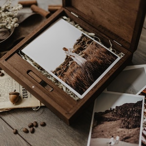 May include: A wooden box with a hinged lid, containing a photograph of a person in a white dress standing in a field of tall grass. The box is open and the photograph is visible. There are other photographs on the table next to the box.