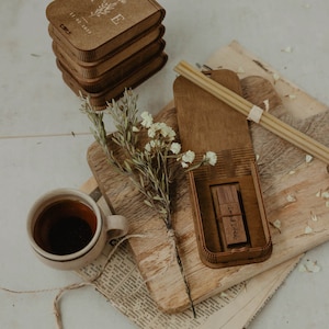 May include: A stack of four rectangular wooden boxes with engraved floral designs and the text 11.02.2025, alongside an open wooden box containing a matching USB drive, a small ceramic cup filled with dark liquid, and a sprig of dried flowers.