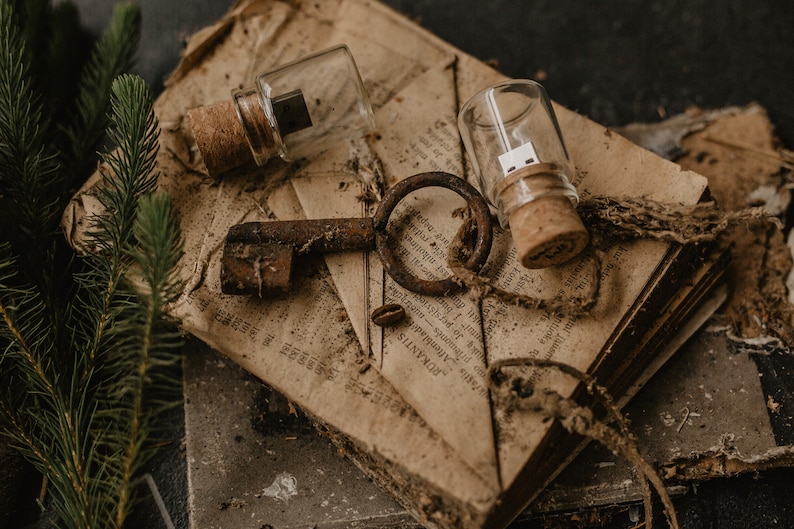 May include: Two glass vials with cork stoppers containing USB drives, an old rusty key, and a piece of twine on top of a stack of old, worn books.