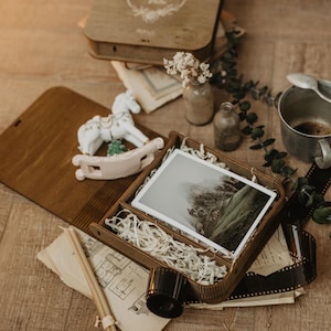 K&ouml;nnte beinhalten: Eine Holzschachtel mit einem Foto einer Berglandschaft, umgeben von einer wei&szlig;en Schaukelpferd-Figur, einem Metallbecher mit L&ouml;ffel, getrockneten Blumen in kleinen Glasflaschen, alten Papierdokumenten und einem Streifen 35mm-Film.