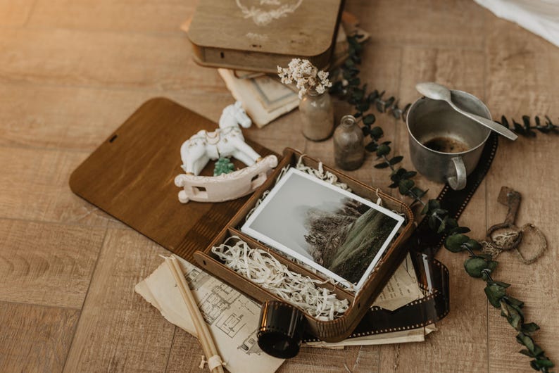 K&ouml;nnte beinhalten: Eine Holzschachtel mit einem Foto einer Berglandschaft, umgeben von einer wei&szlig;en Schaukelpferd-Figur, einem Metallbecher mit L&ouml;ffel, getrockneten Blumen in kleinen Glasflaschen, alten Papierdokumenten und einem Streifen 35mm-Film.