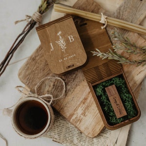 May include: A wooden box with a lid engraved with the initials "J & B" and the date "23.02.2023". The box is open and contains a small wooden USB drive with the text "Daisy & Blake" engraved on it. The USB drive is surrounded by green moss.