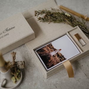 May include: A beige linen box with a photo print and a USB drive inside. The box is engraved with the text "Laura & Antoine 22.07.2023".