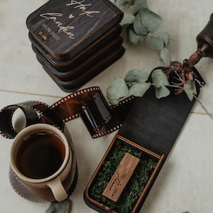 Peut inclure: Une pile de bo&icirc;tes en bois fonc&eacute; avec le texte grav&eacute; "Hayley & Landen 10.08.2022" et une bo&icirc;te ouverte contenant une cl&eacute; USB en bois marqu&eacute;e "Mr. & Mrs. Adams" sur de la mousse verte, avec une tasse en c&eacute;ramique et une pellicule 35mm.