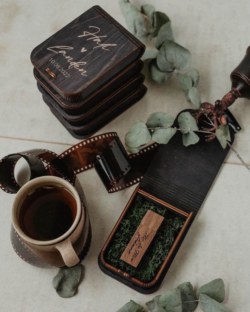 Peut inclure: Une pile de bo&icirc;tes en bois fonc&eacute; avec le texte grav&eacute; "Hayley & Landen 10.08.2022" et une bo&icirc;te ouverte contenant une cl&eacute; USB en bois marqu&eacute;e "Mr. & Mrs. Adams" sur de la mousse verte, avec une tasse en c&eacute;ramique et une pellicule 35mm.