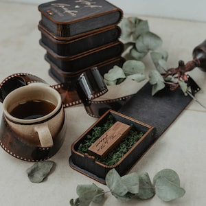 May include: A wooden USB drive with the text "Mr & Mrs Forever" engraved on it. The drive is in a small wooden box with a green moss lining. The box is open and the drive is visible.