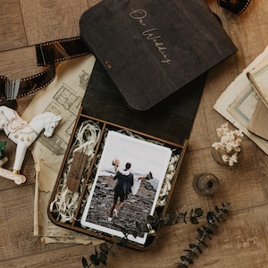 K&ouml;nnte beinhalten: Eine dunkelbraune Holzschachtel mit der Gravur Our Wedding auf dem Deckel. Die ge&ouml;ffnete Schachtel enth&auml;lt ein Foto eines Paares, einen kleinen USB-Stick aus Holz und Papierf&uuml;llmaterial. Die Schachtel ist von Filmstreifen und Dekoartikeln umgeben.
