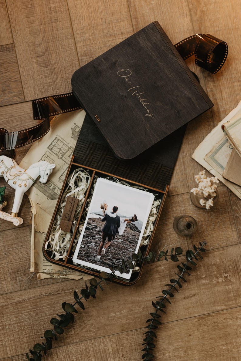 K&ouml;nnte beinhalten: Eine dunkelbraune Holzschachtel mit der Gravur Our Wedding auf dem Deckel. Die ge&ouml;ffnete Schachtel enth&auml;lt ein Foto eines Paares, einen kleinen USB-Stick aus Holz und Papierf&uuml;llmaterial. Die Schachtel ist von Filmstreifen und Dekoartikeln umgeben.