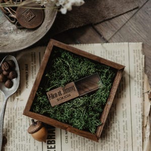 May include: A wooden USB drive and box set, perfect for a wedding or special occasion. The box is filled with green moss and the USB drive is engraved with the names "Alice et Jimmy" and the date "31.08.2024".