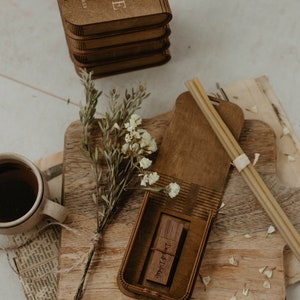 May include: A wooden box with the initials "M & E" and the date "11.12.2023" engraved on the top. The box is open and a wooden USB drive with the text "Dust & Details" is inside. The box is sitting on a wooden surface with a bouquet of white flowers.