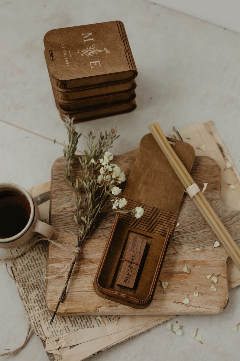 May include: A wooden box with the initials "M & E" and the date "11.12.2023" engraved on the top. The box is open and a wooden USB drive with the text "Dust & Details" is inside. The box is sitting on a wooden surface with a bouquet of white flowers.