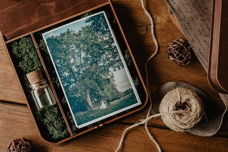 May include: A wooden box with a green moss lining, containing a printed photograph of a couple standing under a large tree, and a small glass bottle with a cork stopper.