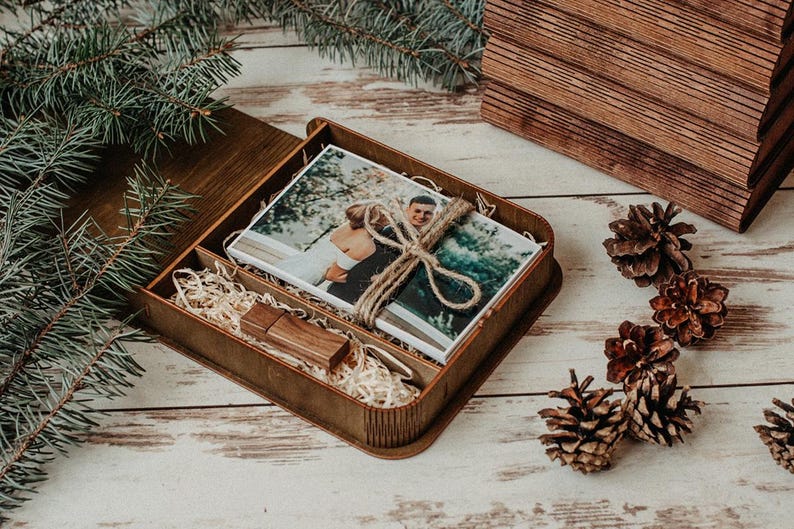 K&ouml;nnte beinhalten: Eine rechteckige Schatulle aus dunklem Holz enth&auml;lt einen Stapel gedruckter Fotos, die mit Jutekordel zusammengebunden sind, sowie einen passenden USB-Stick aus Holz. Das Innere der Box ist mit hellen Holzsp&auml;nen ausgelegt und der Deckel ist offen.