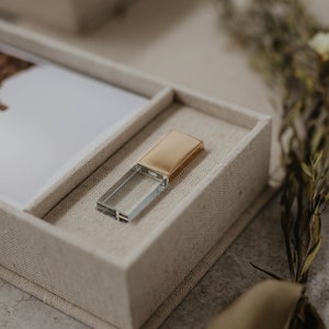 May include: A clear glass USB drive with a gold metal top sits in a beige fabric box.