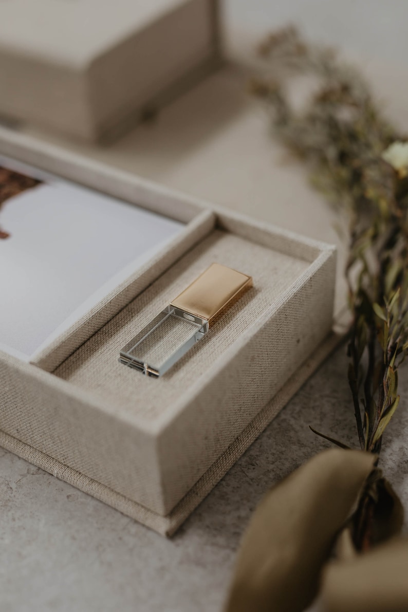 May include: A clear glass USB drive with a gold metal top sits in a beige fabric box.