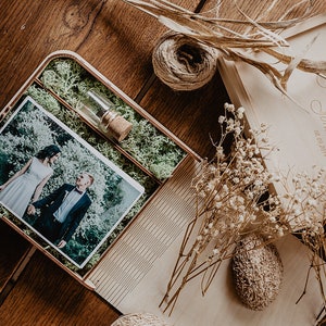 May include: A wooden box with a photo of a couple in a green setting. The box is lined with moss and has a small glass vial with a cork stopper. The box is engraved with the text "Our love story Julia & Johannes 06.02.2017".