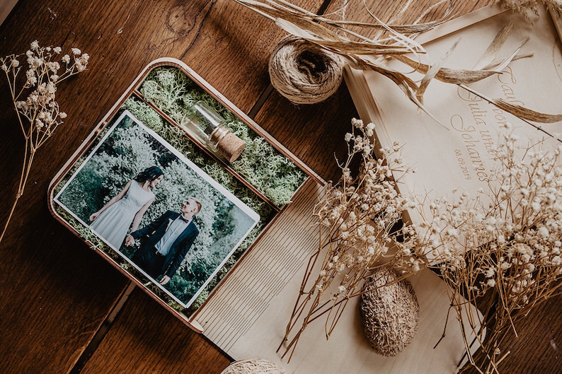 May include: A wooden box with a photo of a couple in a green setting. The box is lined with moss and has a small glass vial with a cork stopper. The box is engraved with the text "Our love story Julia & Johannes 06.02.2017".