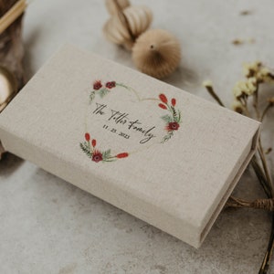 May include: A beige linen box with a heart-shaped wreath design featuring red flowers and green leaves. The box is personalized with the text "The Titter Family" and the date "11.25.2023".