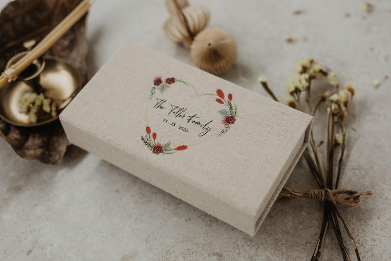 May include: A beige linen box with a heart-shaped wreath design featuring red flowers and green leaves. The box is personalized with the text "The Titter Family" and the date "11.25.2023".