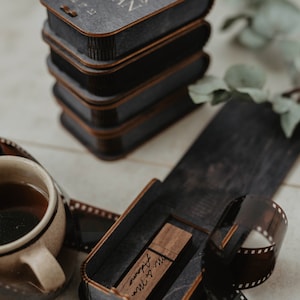 May include: A wooden box with a USB drive inside. The box is engraved with the text "Mr. & Mrs. Johnson" and the date "June 2023". The box is sitting on a table with a roll of film and a cup of coffee.