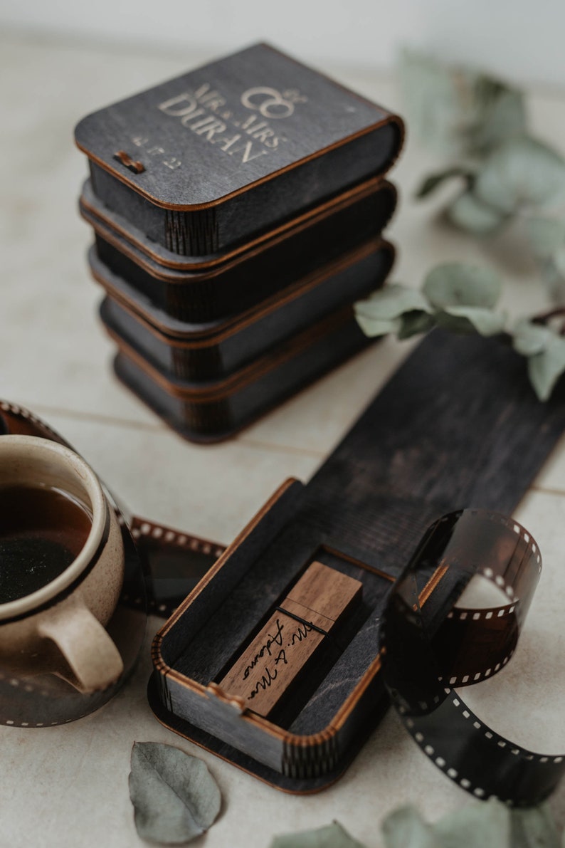 May include: A wooden box with a USB drive inside. The box is engraved with the text "Mr. & Mrs. Johnson" and the date "June 2023". The box is sitting on a table with a roll of film and a cup of coffee.