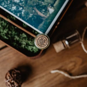 May include: A small glass bottle with a cork top. The cork has a label that reads "The Ground Round". The bottle is sitting on a wooden surface with green moss in the background.